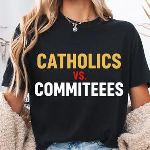 Op de afbeelding: Zwart T-shirt met de tekst "CATHOLICS vs. COMMITTEES" in geel en wit. Het shirt maakt deel uit van de collectie van een kledingwinkel. Het model draagt een blauwe spijkerbroek en een beige vest.