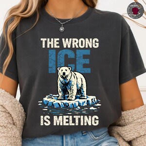 Puede incluir: Camiseta gris oscuro con el texto "THE WRONG ICE IS MELTING". El diseño presenta un oso polar sobre un témpano de hielo derritiéndose, con la palabra "ICE" en letras azules. La camiseta es de cuello redondo.