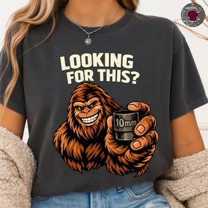 Könnte beinhalten: Dunkelgraues T-Shirt mit einem grinsenden Bigfoot, der eine 10-mm-Steckschlüssel hält. Der Text "LOOKING FOR THIS?" ist über der Grafik aufgedruckt. Das T-Shirt hat einen lässigen Stil.