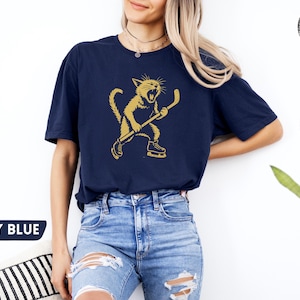 May include: Navy blue t-shirt with a gold illustration of a cat playing hockey. The cat is wearing skates and holding a hockey stick. The text "NAVY BLUE" is in a blue speech bubble in the lower left corner.