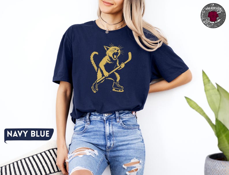 May include: Navy blue t-shirt with a gold illustration of a cat playing hockey. The cat is wearing skates and holding a hockey stick. The text "NAVY BLUE" is in a blue speech bubble in the lower left corner.
