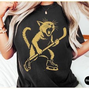 May include: Black t-shirt with a gold illustration of a cat playing hockey. The cat is roaring and wearing ice skates, holding a hockey stick. The shirt has a crew neck.
