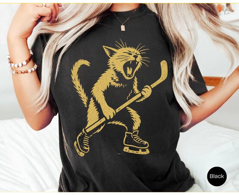 May include: Black t-shirt with a gold illustration of a cat playing hockey. The cat is roaring and wearing ice skates, holding a hockey stick. The shirt has a crew neck.
