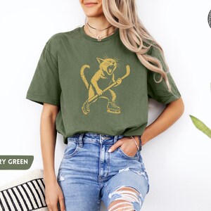 May include: An olive green t-shirt with a gold illustration of a cat playing hockey. The cat is wearing skates and holding a hockey stick. The shirt has a relaxed fit and short sleeves. The text "MILITARY GREEN" is in a green rectangle.