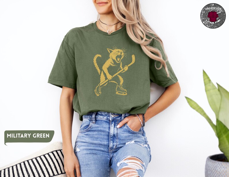 May include: An olive green t-shirt with a gold illustration of a cat playing hockey. The cat is wearing skates and holding a hockey stick. The shirt has a relaxed fit and short sleeves. The text "MILITARY GREEN" is in a green rectangle.