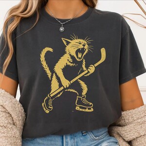 May include: Dark grey t-shirt with a gold illustration of a cat playing hockey. The cat is wearing skates and holding a hockey stick, with its mouth open in a yell. The shirt is a casual style.