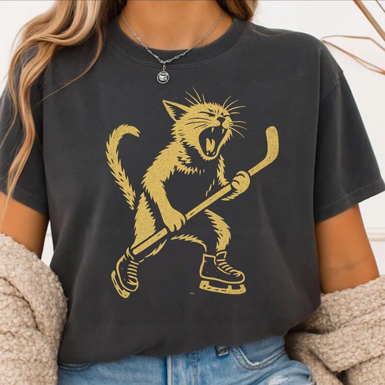 May include: Dark grey t-shirt with a gold illustration of a cat playing hockey. The cat is wearing skates and holding a hockey stick, with its mouth open in a yell. The shirt is a casual style.