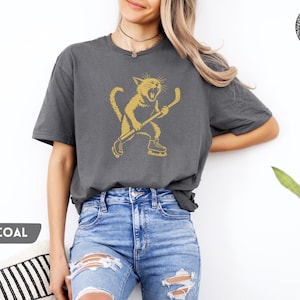 May include: Dark grey t-shirt with a gold illustration of a cat playing hockey. The cat is wearing skates and holding a hockey stick. The word "CHARCOAL" is displayed in a grey box on the bottom left.