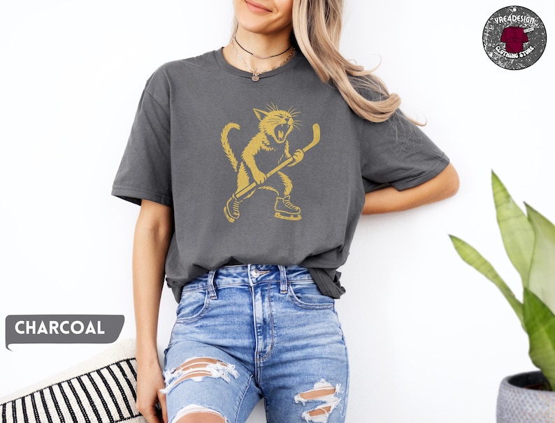 May include: Dark grey t-shirt with a gold illustration of a cat playing hockey. The cat is wearing skates and holding a hockey stick. The word "CHARCOAL" is displayed in a grey box on the bottom left.