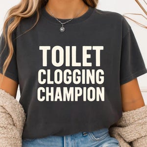 May include: Dark gray t-shirt with the text "TOILET CLOGGING CHAMPION" in large, white capital letters. The shirt has a crew neck and short sleeves. The shirt is made of a soft material.