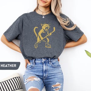 May include: Dark heather grey t-shirt with a gold illustration of a cat playing hockey. The cat is wearing skates and holding a hockey stick. The text "DARK HEATHER" is in a speech bubble.