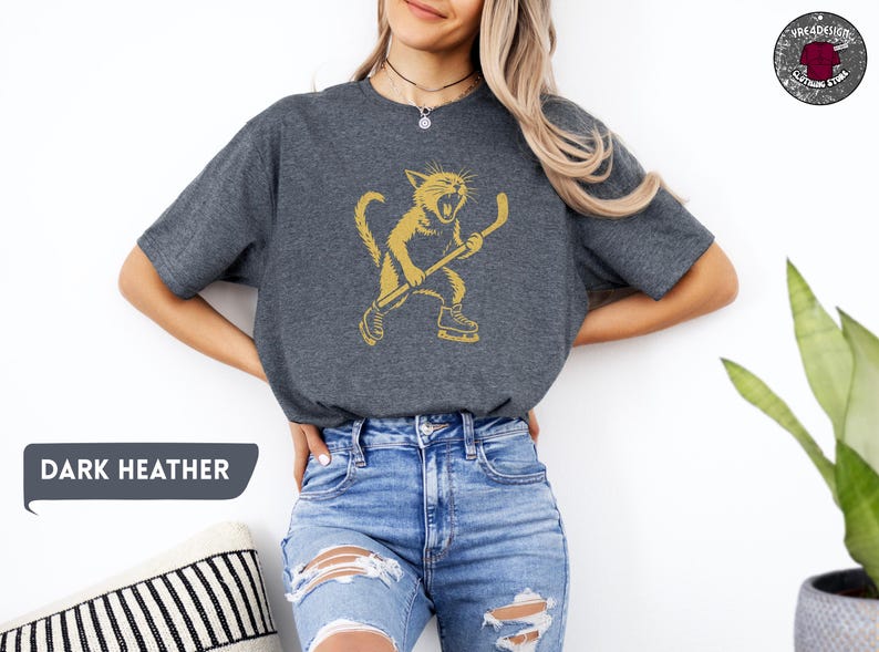 May include: Dark heather grey t-shirt with a gold illustration of a cat playing hockey. The cat is wearing skates and holding a hockey stick. The text "DARK HEATHER" is in a speech bubble.