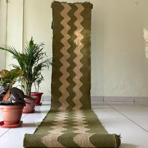 May include: A long, rectangular runner rug featuring a repeating vertical zigzag pattern in olive green and natural beige. The woven textile has a textured surface and frayed edges at the top and bottom ends, displayed in a partially unrolled position.