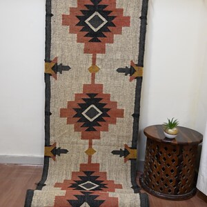 Puede incluir: Una alfombra de pasillo rectangular con un dise&ntilde;o geom&eacute;trico en beige, rojo, negro y amarillo. La alfombra est&aacute; parcialmente enrollada, mostrando su textura y dise&ntilde;o. Cuelga de una barra met&aacute;lica.