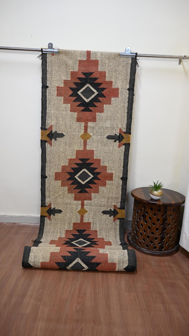 Puede incluir: Una alfombra de pasillo rectangular con un dise&ntilde;o geom&eacute;trico en beige, rojo, negro y amarillo. La alfombra est&aacute; parcialmente enrollada, mostrando su textura y dise&ntilde;o. Cuelga de una barra met&aacute;lica.