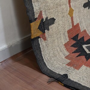 Puede incluir: Una alfombra tejida con un dise&ntilde;o geom&eacute;trico en tonos beige, negro, naranja y amarillo. La alfombra tiene un borde gris oscuro y est&aacute; colocada sobre un suelo de madera. El dise&ntilde;o presenta formas de diamante y estrella.