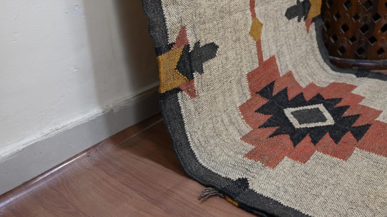 Puede incluir: Una alfombra tejida con un dise&ntilde;o geom&eacute;trico en tonos beige, negro, naranja y amarillo. La alfombra tiene un borde gris oscuro y est&aacute; colocada sobre un suelo de madera. El dise&ntilde;o presenta formas de diamante y estrella.