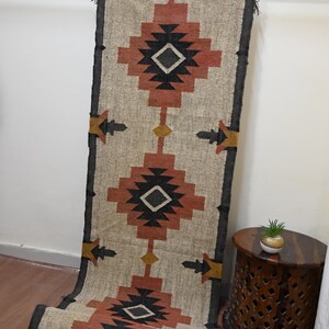 Puede incluir: Una alfombra rectangular beige con un patr&oacute;n geom&eacute;trico en tonos rojos, negros y amarillos. La alfombra est&aacute; colgada de una barra de metal y tiene un borde oscuro. El dise&ntilde;o presenta formas de diamantes y flechas, adecuada para la decoraci&oacute;n del hogar.