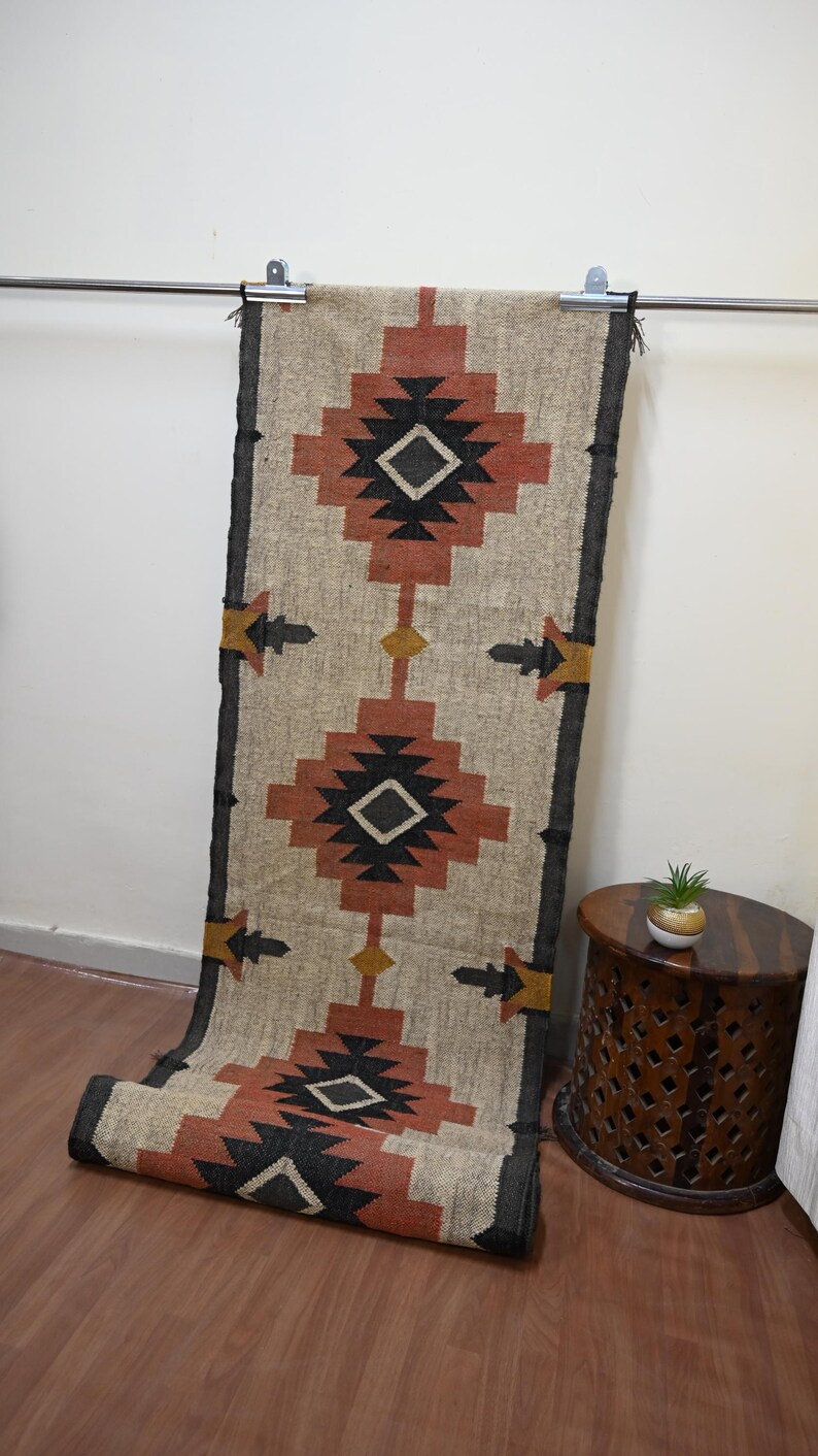 Puede incluir: Una alfombra rectangular beige con un patr&oacute;n geom&eacute;trico en tonos rojos, negros y amarillos. La alfombra est&aacute; colgada de una barra de metal y tiene un borde oscuro. El dise&ntilde;o presenta formas de diamantes y flechas, adecuada para la decoraci&oacute;n del hogar.