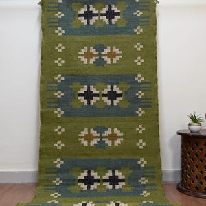 以下が含まれることがあります： 幾何学模様の緑と青のランナーラグ。ラグは、黒と白の十字とひし形の繰り返しモチーフが特徴です。ラグは織物でできており、金属製の棒から吊り下げて展示されています。