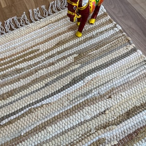 May include: A striped, handwoven rug in shades of beige, brown, and white. A decorative wooden horse figurine, painted in red, green, yellow, and blue, is placed on the rug. The rug features a fringed edge.