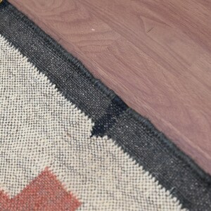 Puede incluir: Primer plano de una alfombra tejida con un patr&oacute;n geom&eacute;trico. La alfombra presenta un fondo crema con zigzags rojos, negros y amarillos. Un borde gris oscuro enmarca la alfombra, que est&aacute; colocada sobre una superficie de madera.