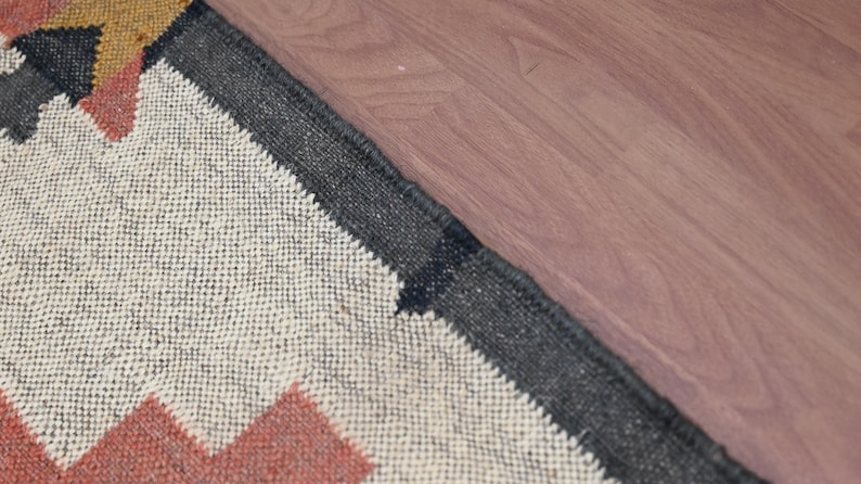 Puede incluir: Primer plano de una alfombra tejida con un patr&oacute;n geom&eacute;trico. La alfombra presenta un fondo crema con zigzags rojos, negros y amarillos. Un borde gris oscuro enmarca la alfombra, que est&aacute; colocada sobre una superficie de madera.