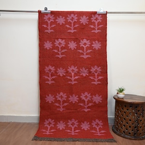 Puede incluir: Una alfombra rectangular roja con un estampado floral en un tono más claro de rojo. La alfombra cuelga de una barra de metal. La alfombra tiene un borde con flecos en la parte inferior. La alfombra está hecha de un material tejido.