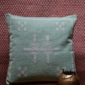 May include: A square, pale green cushion featuring white geometric designs. The cushion has a textured finish and a white border. The background is a red and white patterned fabric.