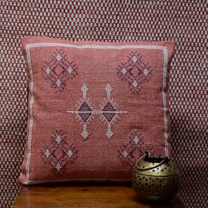 May include: A square, coral-colored decorative pillow with embroidered geometric patterns in white and purple. The pillow has a white border and is placed on a wooden surface. A small, gold-colored metal lantern sits nearby.