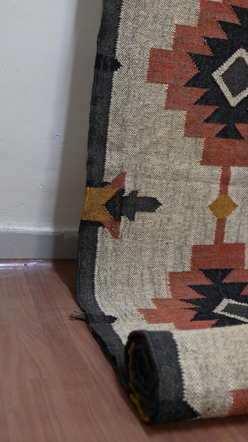 Puede incluir: Una alfombra enrollada con un patr&oacute;n geom&eacute;trico beige, negro y &oacute;xido. El dise&ntilde;o incluye formas repetidas en negro, &oacute;xido y amarillo. La alfombra est&aacute; hecha de material tejido y est&aacute; parcialmente enrollada, mostrando la parte inferior.