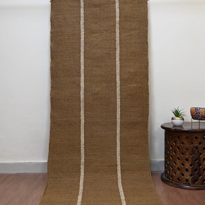 Può includere: Tappeto marrone tessuto con due strisce verticali bianco sporco. Il tappeto è appeso a una barra di metallo con clip. Il tappeto è arrotolato in basso. Il tappeto è fatto di fibra naturale.