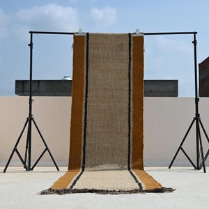 May include: A long rectangular table runner made of natural tan woven jute fabric. The runner features two thick mustard yellow vertical stripes along the sides, each bordered by a thin black line. The bottom edge has a frayed, fringed finish.
