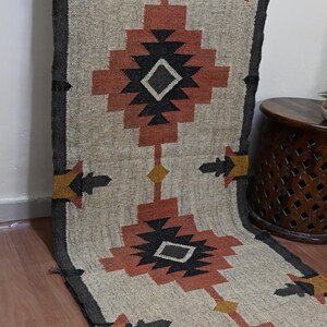 Puede incluir: Una alfombra de pasillo beige y negra con un patr&oacute;n geom&eacute;trico. La alfombra presenta formas repetidas de diamantes y flechas en negro, &oacute;xido y amarillo. La alfombra est&aacute; hecha de un material tejido y tiene un borde gris oscuro.