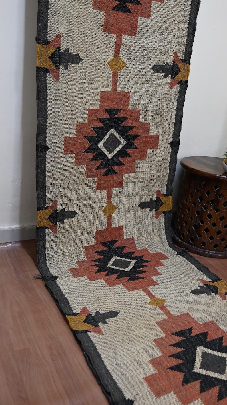 Puede incluir: Una alfombra de pasillo beige y negra con un patr&oacute;n geom&eacute;trico. La alfombra presenta formas repetidas de diamantes y flechas en negro, &oacute;xido y amarillo. La alfombra est&aacute; hecha de un material tejido y tiene un borde gris oscuro.