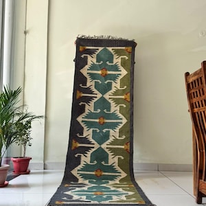 May include: A long rectangular runner rug featuring a repeating geometric pattern in teal, olive green, and cream. The design includes small yellow accents at the center of each motif, finished with dark gray fringed edges on the top and bottom ends.