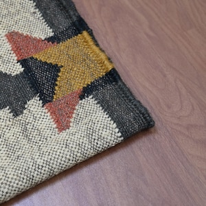 Puede incluir: Primer plano de una alfombra tejida con un dise&ntilde;o geom&eacute;trico. La alfombra presenta un fondo color crema con una forma de flecha gris oscuro y un patr&oacute;n de estrella amarilla y roja. La alfombra est&aacute; colocada sobre un suelo de madera.