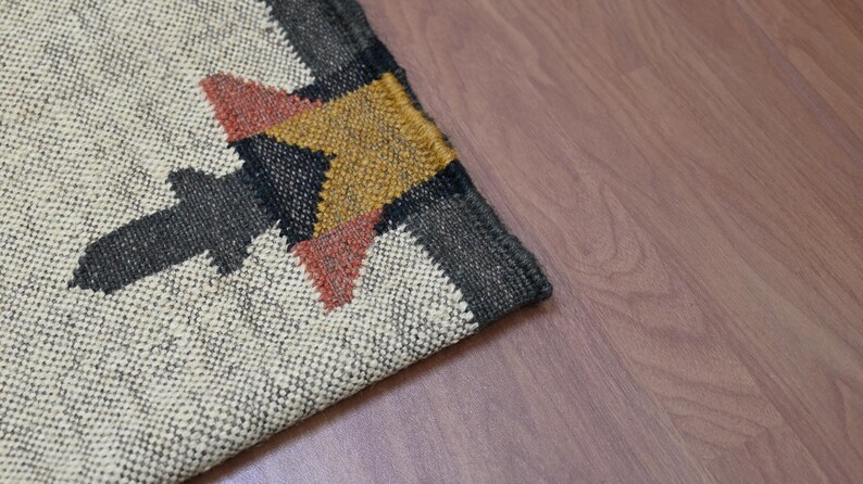 Puede incluir: Primer plano de una alfombra tejida con un dise&ntilde;o geom&eacute;trico. La alfombra presenta un fondo color crema con una forma de flecha gris oscuro y un patr&oacute;n de estrella amarilla y roja. La alfombra est&aacute; colocada sobre un suelo de madera.