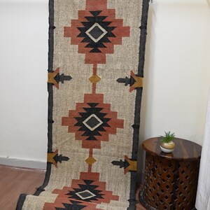 Puede incluir: Una alfombra de pasillo rectangular, beige y color &oacute;xido con un patr&oacute;n geom&eacute;trico. La alfombra presenta formas de diamantes y flechas en negro, &oacute;xido y dorado. La alfombra cuelga de una barra de metal.
