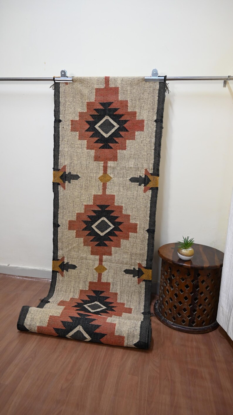 Puede incluir: Una alfombra de pasillo rectangular, beige y color &oacute;xido con un patr&oacute;n geom&eacute;trico. La alfombra presenta formas de diamantes y flechas en negro, &oacute;xido y dorado. La alfombra cuelga de una barra de metal.
