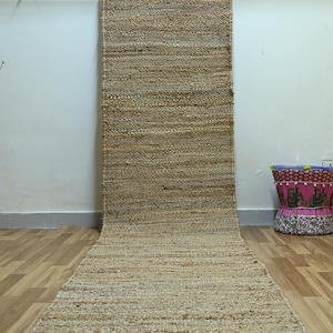 Puede incluir: Una alfombra de pasillo larga y rectangular hecha de fibras naturales trenzadas de color marrón claro. La alfombra presenta una textura trenzada apretada y uniforme en toda su longitud, extendiéndose desde el suelo hasta una barra metálica.