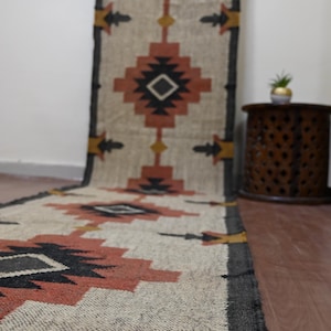 Puede incluir: Una alfombra corredora tejida con un patr&oacute;n geom&eacute;trico en tonos beige, negro, naranja y amarillo. La alfombra presenta formas de diamante y dise&ntilde;os en forma de flecha, adecuada para un pasillo o entrada.