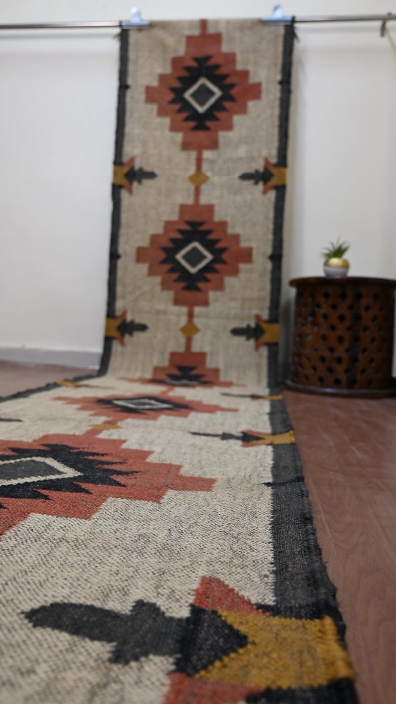 Puede incluir: Una alfombra corredora tejida con un patr&oacute;n geom&eacute;trico en tonos beige, negro, naranja y amarillo. La alfombra presenta formas de diamante y dise&ntilde;os en forma de flecha, adecuada para un pasillo o entrada.