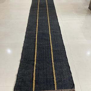 May include: A long, rectangular runner rug featuring a dark charcoal gray woven texture. The rug is accented by two parallel vertical mustard yellow stripes running the length of the piece. The ends are finished with short, light brown fringe.