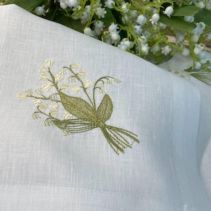 May include: White linen fabric with an embroidered bouquet of lily of the valley flowers in shades of green and cream. The embroidery is detailed and delicate, with a focus on the floral design. A sprig of lily of the valley flowers is in the background.
