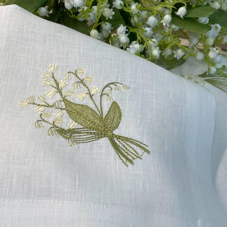 May include: White linen fabric with an embroidered bouquet of lily of the valley flowers in shades of green and cream. The embroidery is detailed and delicate, with a focus on the floral design. A sprig of lily of the valley flowers is in the background.