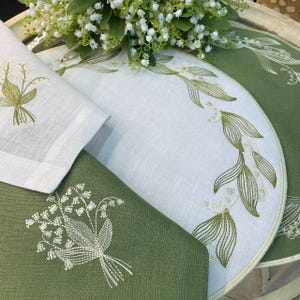 May include: White and green embroidered linen placemats and napkins. The placemats have a half-circle shape with a floral design. The napkins and placemats feature a lily of the valley embroidery. A bouquet of white flowers is in the background.