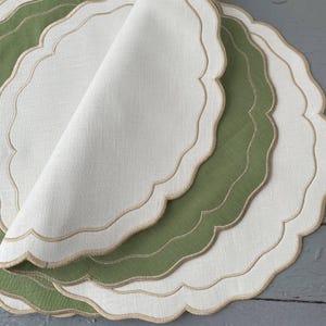 May include: A stack of scalloped-edge placemats in white and green. The placemats have a layered design with a beige trim. The fabric appears to be linen or a similar natural material, with a slightly textured surface.