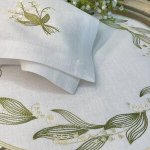 May include: White linen table linens with embroidered lily of the valley design. The set includes a folded napkin and a placemat with green and cream-colored floral embroidery. The linens are arranged on a round tray.