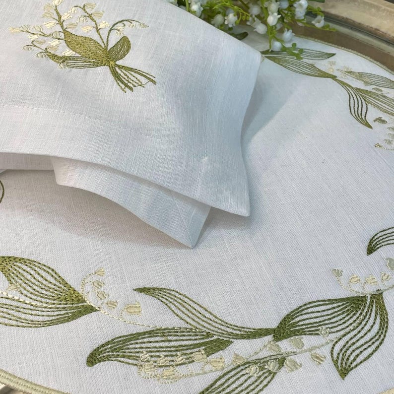 May include: White linen table linens with embroidered lily of the valley design. The set includes a folded napkin and a placemat with green and cream-colored floral embroidery. The linens are arranged on a round tray.